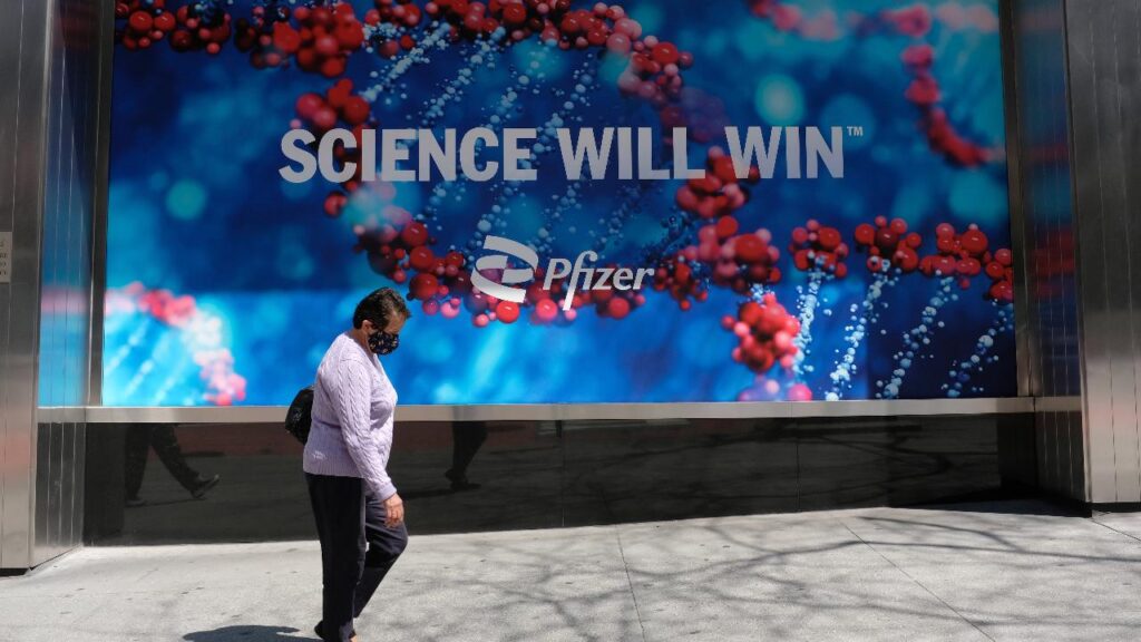 The science will win New York City, NY, USA - 20 aprile 2021: Persone in maschera che camminano accanto ai cartelli fuori dal Pfizer Building sulla 42nd Street. Pfizer ha distribuito il suo vaccino COVID-19.